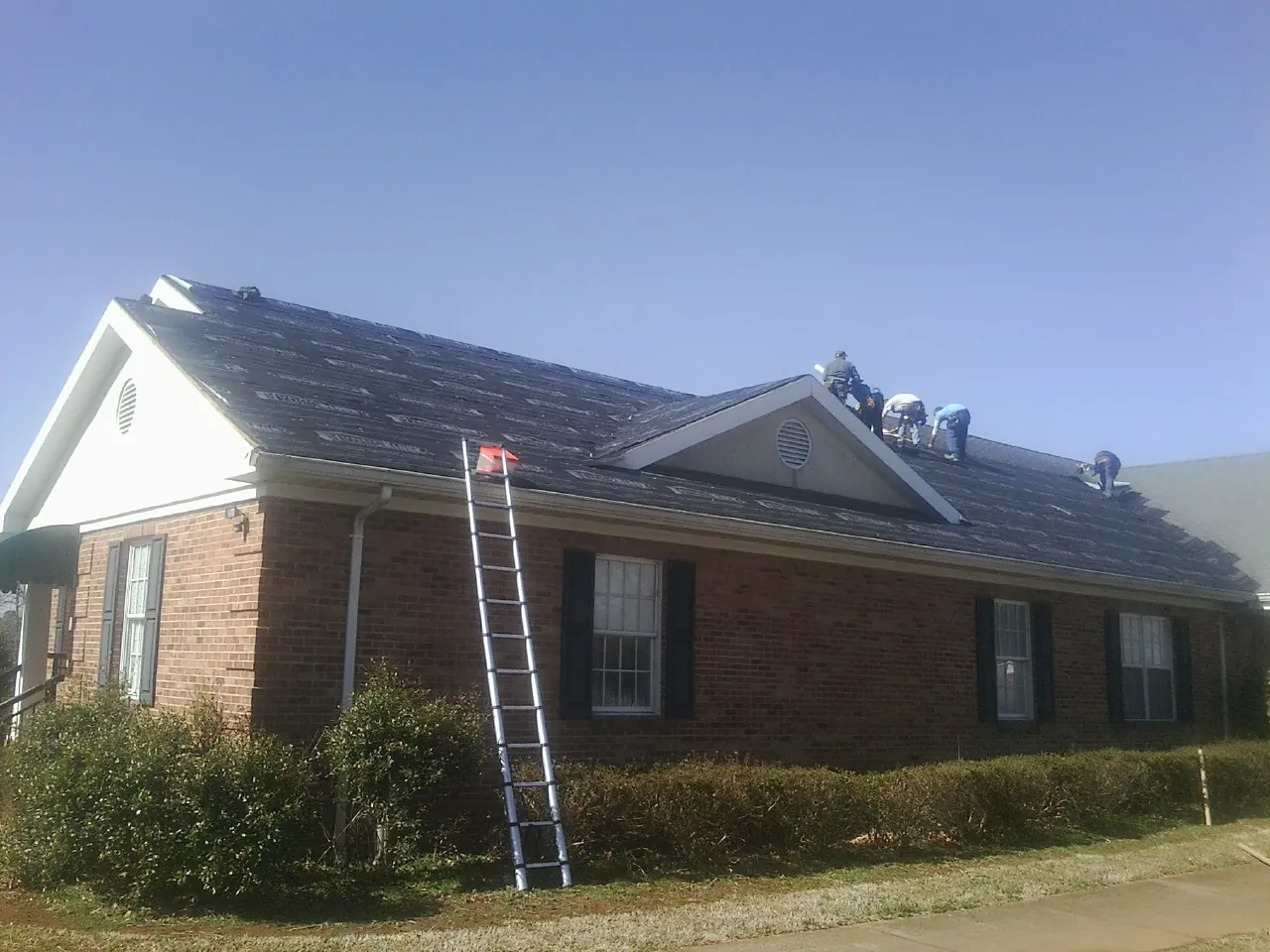 Crew tearing shingles off a brick ranch roof with a ladder set — Metal Roof Repair work by our crew in Hysham