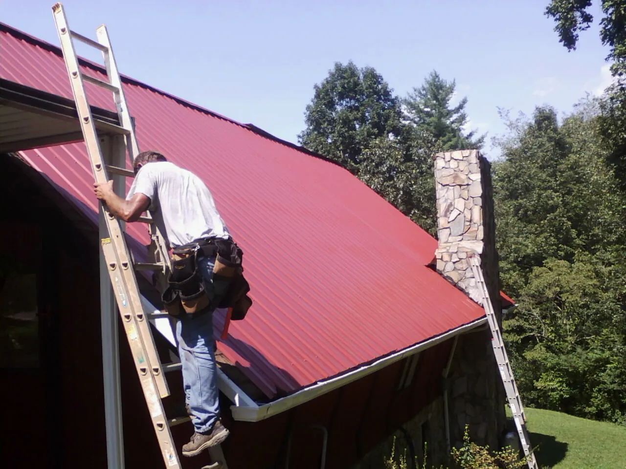 Roofer on a ladder beside a red metal roof and stone chimney — Soffit & Fascia Repair by Roofing Brothers in Hysham